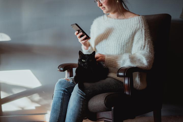 pet owner in white sweater sitting on chair while using phone with black cat sits on lap