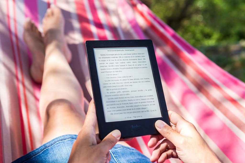 white person laying on a pink red and white hammock wearing a denim skirt while holding a black ereader with a screen showing a book page