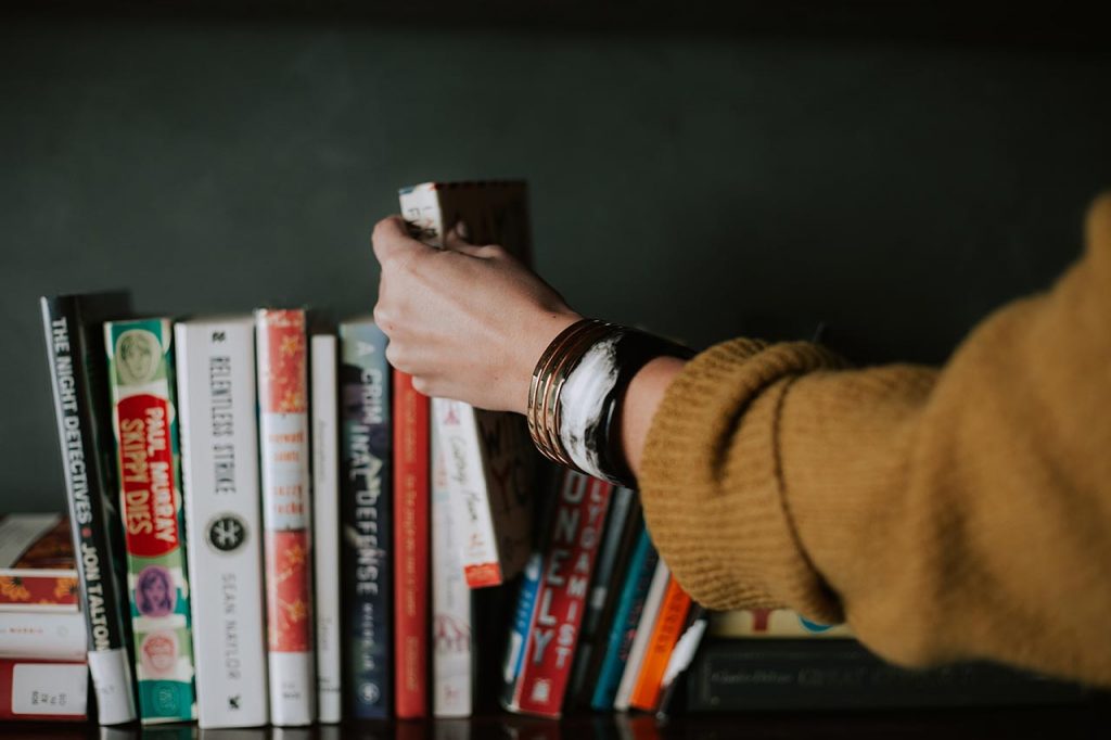white person wearing a yellow sweater and bracelets grabbing a white and red book from a row of different books with a dark gray green background