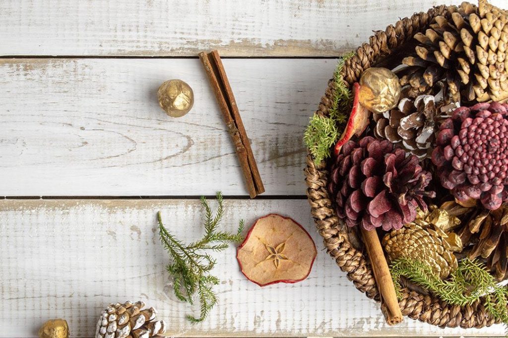 wicker bowl of brown and red pine cones cinnamon sticks dried apple slices and conifer leaves sitting on a white wooden table with similar items scattered around