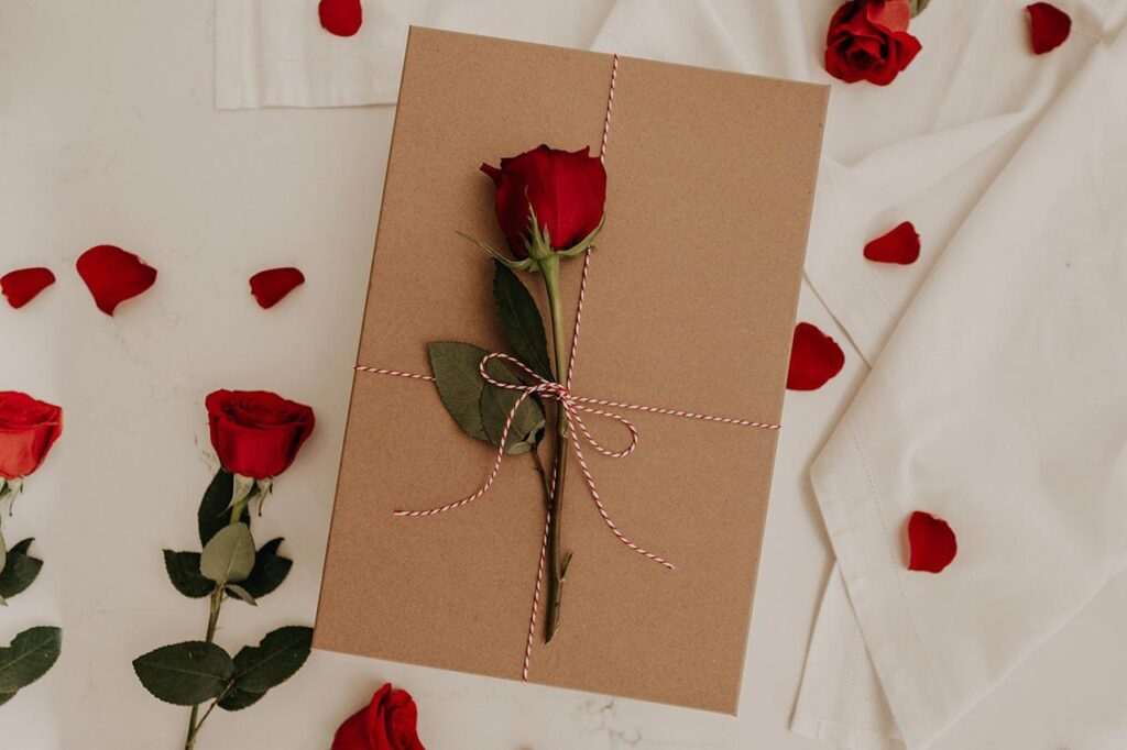 a rose tied with striped string on brown paper that is atop a white sheet, surrounded by rose petals