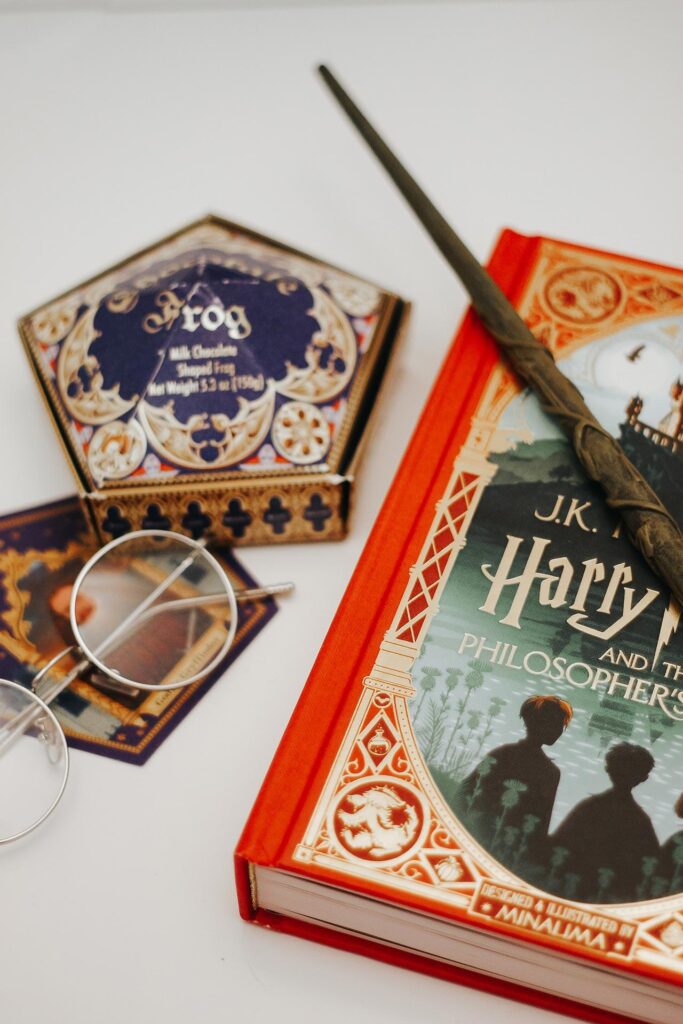 A wand sits atop a “Harry Potter” book with Harry’s glasses, a Chocolate Frog box, and a Chocolate Frog card surrounding it